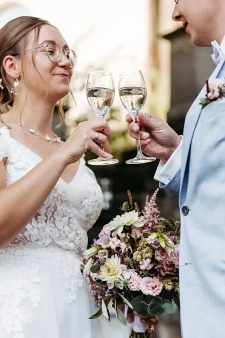 Eine Braut im weißen Spitzenkleid hält ein Glas Champagner und einen Blumenstrauß in der Hand und stößt mit ihrem Bräutigam im hellblauen Anzug an. Zwischen den zarten Blüten lächeln sie sich sanft an. Eingefangen von Melanie Kunert Fotografie, ist dieser Moment in Marcolinis Welt pure Magie.