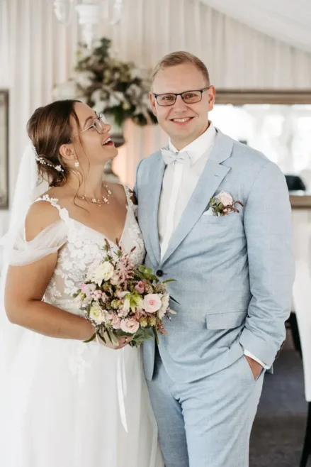 Eine Braut im weißen Kleid schaut den Bräutigam im hellblauen Anzug lächelnd an. Ein Hochzeitsfotograf in Marcolinis Welt hat das Bild wunderschön festgehalten. Sie hält einen Blumenstrauß in der Hand, und beide lächeln strahlend. Sie stehen im Innenbereich, im Hintergrund ein beiger Vorhang und ein Spiegel.