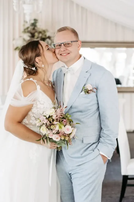 Eine Braut im weißen Kleid küsst den Bräutigam im hellblauen Anzug auf die Wange. Ein Hochzeitsfotograf in Marcolinis Welt hat das Bild wunderschön festgehalten. Sie hält einen Blumenstrauß in der Hand, und beide lächeln strahlend. Sie stehen im Innenbereich, im Hintergrund ein beiger Vorhang und ein Spiegel.