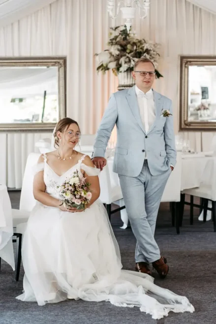 Eine Braut im weißen Kleid hält einen Blumenstrauß in der Hand, während sie neben ihrem Bräutigam im hellblauen Anzug sitzt. Aufgenommen von Melanie Kunert Fotografie posieren sie elegant in einem dekorierten Raum mit weißen Vorhängen und gerahmten Spiegeln.