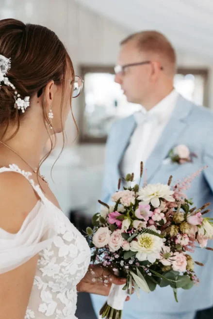 Eine Braut in einem weißen Spitzenkleid hält einen Strauß rosa, weißer und violetter Blumen in den Händen und blickt ihren Bräutigam im hellblauen Anzug an. Die Inneneinrichtung, aufgenommen von einem Hochzeitsfotografen in Marcolinis Welt, besticht durch sanftes Licht und neutrales Dekor.