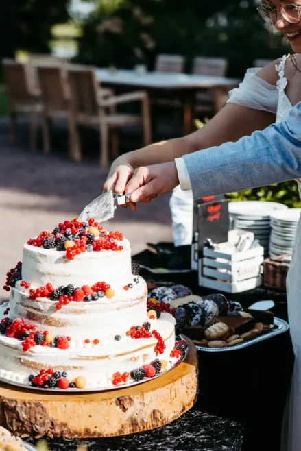 Braut und Bräutigam schneiden eine dreistöckige Hochzeitstorte an, die mit roten und schwarzen Beeren verziert ist. Ein Hochzeitsfotograf in Marcolinis Welt hat die Torte auf einem Holzständer, umgeben von verschiedenen Gebäckstücken, vor einer eleganten Außenkulisse mit Tischen und Stühlen festgehalten.