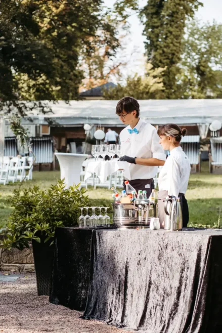 Zwei Kellner in weißen Hemden und Fliege stehen hinter einem mit einem schwarzen Tuch bedeckten Tisch im Freien und bereiten Weingläser und Flaschen in einem Eiskübel vor. Dekorierte Tische und eine Gartenkulisse bilden die perfekte Kulisse, ideal für die Erinnerungsfotos mit einem Hochzeitsfotografen in Marcolinis Welt.