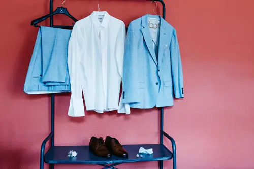 Ein hellblaues Sakko und eine hellblaue Anzughose, ein weißes Hemd, braune Schuhe, eine blaue Fliege und Manschettenknöpfe werden elegant auf einem blauen Kleiderständer vor einer roten Wand präsentiert. Dieses Ensemble eignet sich ideal für Fotos eines Hochzeitsfotografen im Hotel Rothenburger Hof.