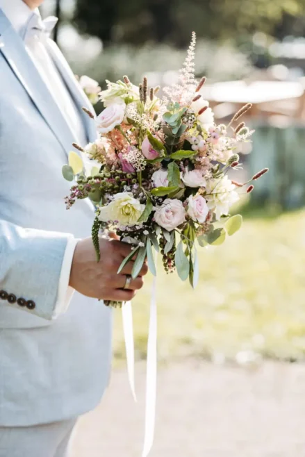 Eine Person im hellgrauen Anzug hält einen Strauß pastellfarbener Blumen in der Hand. Ihr leuchtendes Arrangement hebt sich vor dem unscharfen Hintergrund hervor. Das durchscheinende Sonnenlicht verstärkt die ruhige Schönheit dieser Szene im Freien – eine perfekte Aufnahme für jeden Hochzeitsfotografen in Marcolinis Welt.