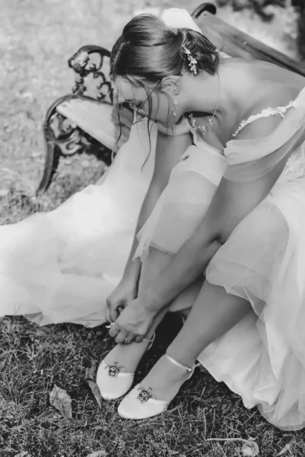 Eine Braut in einem fließenden weißen Kleid sitzt anmutig auf einer Vintage-Bank im Freien und rückt den Riemen ihres Schuhs zurecht. Ihr mit einem Blumenschmuck geschmücktes Haar strahlt Eleganz aus. Eingefangen von einem Hochzeitsfotografen in Marcolinis Welt, strahlt die Schwarz-Weiß-Szene klassische Zeitlosigkeit aus.