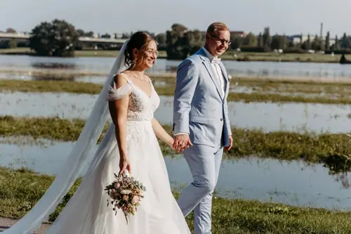Eine Braut im weißen Kleid mit Blumenstrauß und ein Bräutigam im hellblauen Anzug schlendern Hand in Hand am Wasser entlang. Ein Hochzeitsfotograf in Marcolinis Welt fotografierte sie lächelnd unter klarem Himmel. Grünes Grün und Bäume in der Ferne runden die malerische Szene ab.