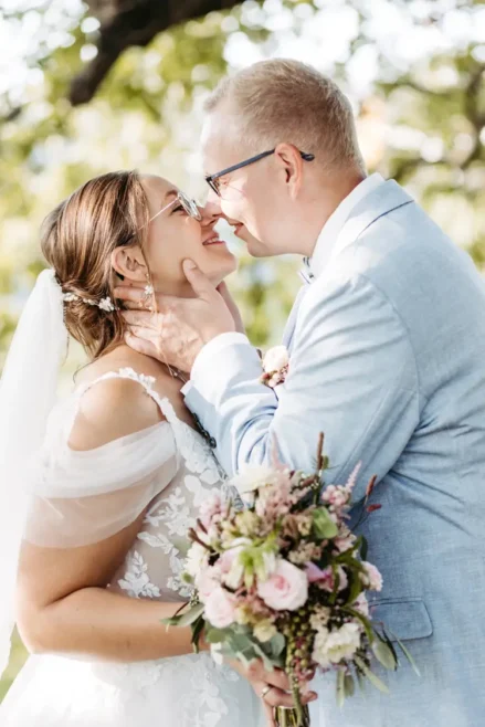 Ein talentierter Hochzeitsfotograf in Marcolinis Welt hält ein Paar fest, das sich an seinem Hochzeitstag liebevoll umarmt. Die Braut in einem floralen Spitzenkleid hält einen Strauß rosa und weißer Blumen in der Hand, während sie sich unter dem sonnendurchfluteten Baum herzlich anlächeln. Der Bräutigam trägt einen hellblauen Anzug.
