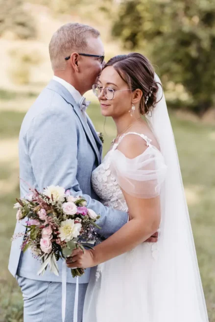 Braut und Bräutigam stehen sich umarmend in einer sonnigen Außenkulisse gegenüber, aufgenommen von einem Hochzeitsfotografen aus Dresden in der Nähe von Marcolinis Welt . Die Braut trägt ein weißes Kleid mit langem Schleier, der Bräutigam einen hellgrauen Anzug. Umgeben von Gras und Bäumen entsteht eine friedliche Atmosphäre.