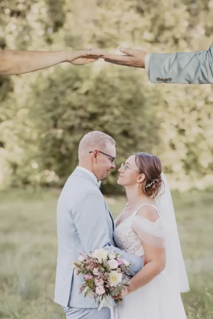Braut und Bräutigam, aufgenommen von einem Hochzeitsfotografen in der Nähe von Marcolinis Welt, blicken sich liebevoll in die Augen, umgeben von üppigem Grün. Die Braut hält einen Blumenstrauß und trägt einen Schleier; der Bräutigam trägt einen hellen Anzug, und ihre Hände berühren sich über ihnen.