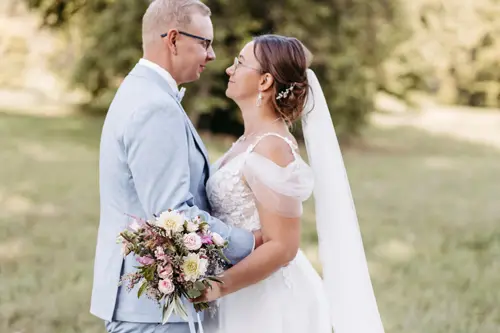 Braut und Bräutigam stehen sich Händchen haltend in einer sonnigen Außenkulisse gegenüber, aufgenommen von einem Hochzeitsfotografen aus Dresden. Die Braut trägt ein weißes Kleid mit langem Schleier, der Bräutigam einen hellgrauen Anzug. Umgeben von Gras und Bäumen entsteht eine friedliche Atmosphäre.