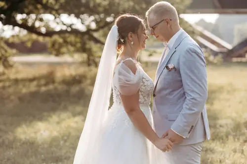 Braut und Bräutigam stehen sich Händchen haltend in einer sonnigen Außenkulisse gegenüber, aufgenommen von einem Hochzeitsfotografen aus Dresden. Die Braut trägt ein weißes Kleid mit langem Schleier, der Bräutigam einen hellgrauen Anzug. Umgeben von Gras und Bäumen entsteht eine friedliche Atmosphäre.