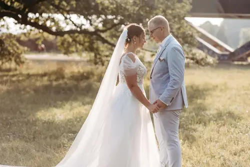 Braut und Bräutigam stehen sich Händchen haltend in einer sonnigen Außenkulisse gegenüber, aufgenommen von einem Hochzeitsfotografen aus Dresden. Die Braut trägt ein weißes Kleid mit langem Schleier, der Bräutigam einen hellgrauen Anzug. Umgeben von Gras und Bäumen entsteht eine friedliche Atmosphäre.