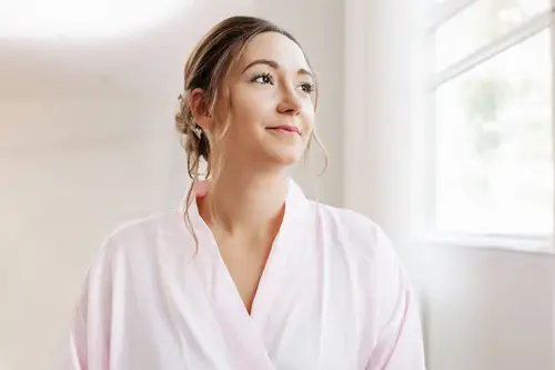 Eine Frau in einem hellrosa Morgenmantel lächelt sanft und blickt aus einem sonnenbeschienenen Fenster. Ihr Haar ist hochgesteckt, wobei lose Strähnen ihr Gesicht umrahmen und die gelassene Eleganz verkörpern. Der Hintergrund ist hell und sanft verschwommen.