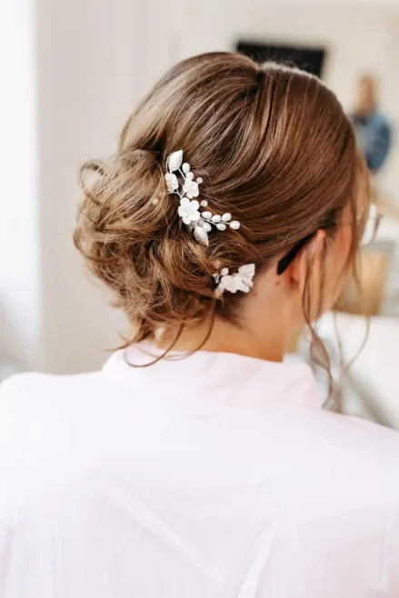Eine elegante Hochsteckfrisur mit lockeren Locken ist mit dekorativen weißen Blumennadeln verziert, perfekt eingefangen von einem Hochzeitsfotografen. Sie trägt ein hellrosa Oberteil, während der sanft verschwommene Hintergrund ihre exquisite Frisur in den Mittelpunkt rückt.