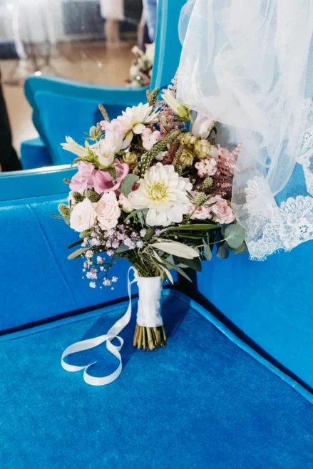 Ein Strauß weißer und rosa Blumen, akzentuiert mit Grün, ist mit einem weißen Band umwickelt und steht vor einem blauen Sitz. Zarte Spitze drapiert elegant daneben, während ein Spiegel einen Teil des Arrangements reflektiert – eine perfekte Szene, eingefangen von einem Hochzeitsfotografen in Hotel Rothenburger Hof.