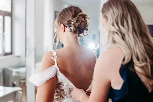 Eine Braut in einem weißen Spitzenkleid wird von einer Frau in Marineblau unterstützt, die den Rücken ihres Kleides zurechtrückt. Ihr Haar, elegant mit Blumenschmuck gestylt, fängt das natürliche Licht ein, das in den hellen Raum strömt – ein Moment, perfekt eingefangen vom Hochzeitsfotografen in Marcolinis Welt.