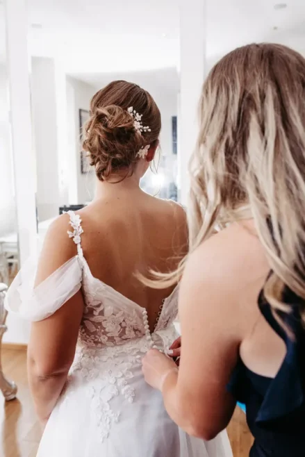 Eine Braut mit einer aufwendigen Hochsteckfrisur und einer Blumenhaarnadel steht da, während eine Frau in einem dunklen Kleid ihr beim Zuknöpfen ihres Spitzenkleides in einem hell erleuchteten Raum hilft.