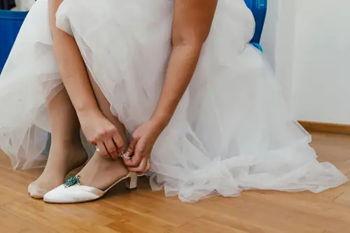 Eine Braut in einem weißen Kleid rückt ihren Absatzschuh mit einer Schmuckbrosche zurecht, während sie auf dem Holzboden sitzt. Ihr anderer Fuß ist von hauchdünnen Strümpfen bedeckt, während die Schichten des Kleides sie elegant umhüllen.