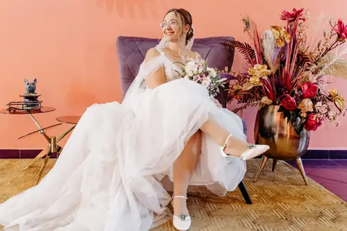 Eine lächelnde Braut in weißem Kleid und Brille sitzt auf einem lila Stuhl und hält einen Blumenstrauß in der Hand. Der Hintergrund zeigt eine pfirsichfarbene Wand und ein farbenfrohes Blumenarrangement in einer Vase. Neben ihr, auf einem kleinen Tisch, steht eine dekorative Katzenfigur – eine perfekte Szene für jeden Hochzeitsfotografen.