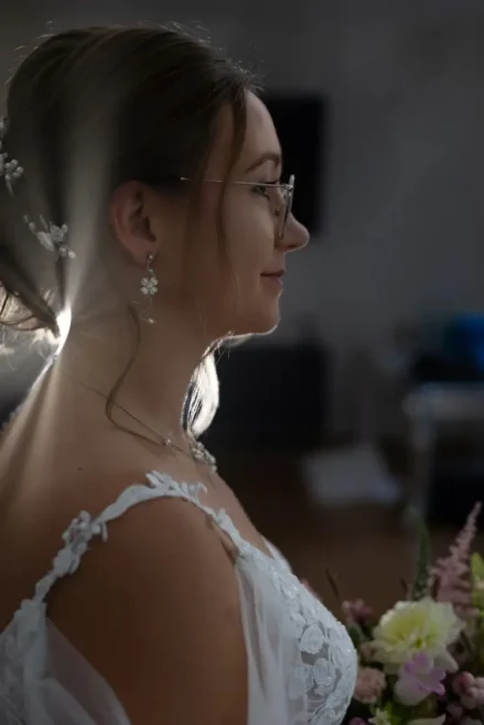Eine Frau im Brautkleid, geschmückt mit floralem Haarschmuck und Brille, steht im Profil und hält einen Blumenstrauß. Sonnenlicht umgibt ihren nachdenklichen Gesichtsausdruck wie ein Heiligenschein. Dieser ätherische Moment, eingefangen von einem Hochzeitsfotografen in Marcolinis Welt, zeichnet sich durch einen sanft verschwommenen Hintergrund aus.