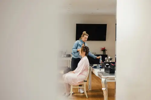 Eine Frau im rosa Bademantel sitzt auf einem Hocker, während eine andere ihr Haar frisiert und sich auf einen Moment vorbereitet, den ein Hochzeitsfotograf eingefangen hat. Die Szene wird teilweise durch einen Türrahmen, Friseurutensilien auf dem Tisch und einen Fernseher an der Wand im Hintergrund verdeckt.
