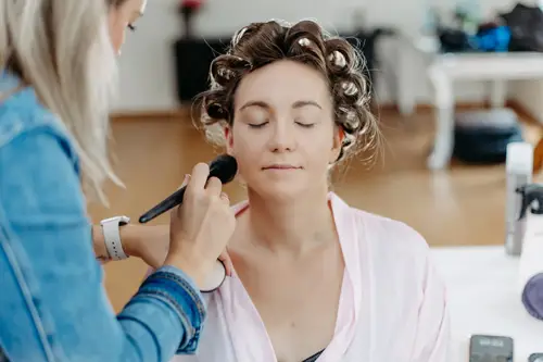 In einem hell erleuchteten Raum voller Kosmetikprodukte wird eine Person von einem Visagisten kunstvoll geschminkt. Die Augen sind geschlossen, die Haare auf Lockenwicklern fixiert. So entsteht ein heiterer Moment, der an die Arbeit eines Hochzeitsfotografen erinnert.