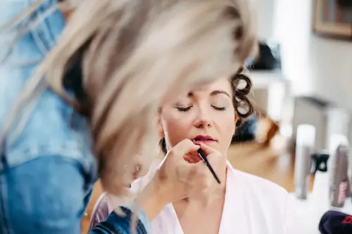 In einem hell erleuchteten Raum voller Kosmetikprodukte wird eine Person von einem Visagisten kunstvoll geschminkt. Die Augen sind geschlossen, die Haare auf Lockenwicklern fixiert. So entsteht ein heiterer Moment, der an die Arbeit eines Hochzeitsfotografen erinnert.