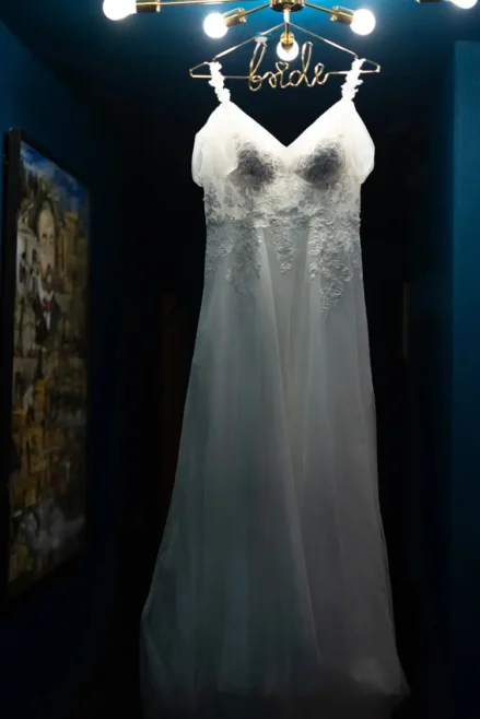 Ein weißes Brautkleid mit der Aufschrift „Braut“ hängt elegant in einem schmalen Flur mit leuchtend blauen Wänden, eingefangen von einem Hochzeitsfotografen in Marcolinis Welt. Das gedämpfte Licht und die gerahmten Bilder bilden einen künstlerischen Kontrast zu dieser bezaubernden Szene.