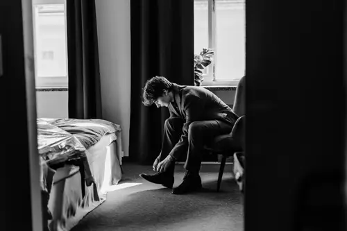 Auf einem Schwarz-Weiß-Bild, aufgenommen von einem Hochzeitsfotografen , sitzt ein Mann im Anzug auf einem Stuhl und bindet seine Schnürsenkel. Sonnenlicht fällt durch die Vorhänge des Bettzimmers und beleuchtet seine nachdenkliche Haltung.