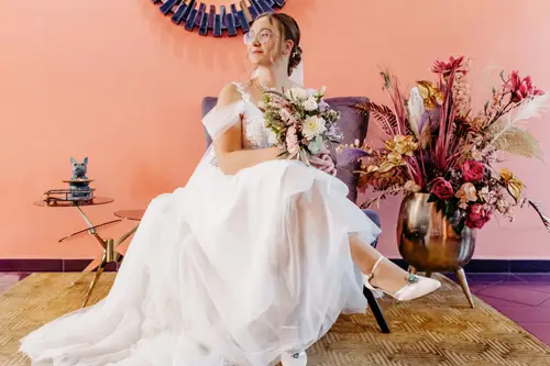 Eine Braut in einem weißen Kleid hält ihren Brautstrauß in der Hand, während sie auf einem lila Stuhl sitzt. Elegant blickt sie zur Seite, eingerahmt von einem pfirsichfarbenen Hintergrund mit einem Sonnenspiegel. Die Kulisse strahlt Anmut aus, ganz wie die raffinierte Kunstfertigkeit des Hochzeitsfotografen im Hotel Rothenburger Hof, der solche Momente einfängt.