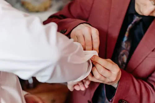 Vor dem unscharfen Hintergrund richtet eine Person in einer roten Jacke vorsichtig die Manschette eines weißen Hemdes, das eine andere Person trägt, und fängt damit einen intimen Moment ein, den jeder Hochzeitsfotograf in Ehren halten würde.
