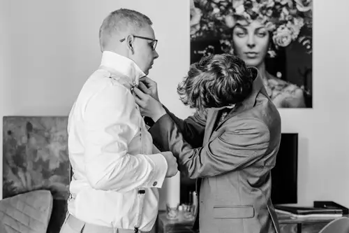 In einer Schwarz-Weiß-Szene fotografiert der Hochzeitsfotograf zwei Männer in formeller Kleidung. Einer trägt ein frisches weißes Hemd und der andere Herr im Anzug hilft ihm beim Zurechtrücken seines Kragens. Hinter ihnen hängt das Porträt einer Frau mit Blumenkopfschmuck.