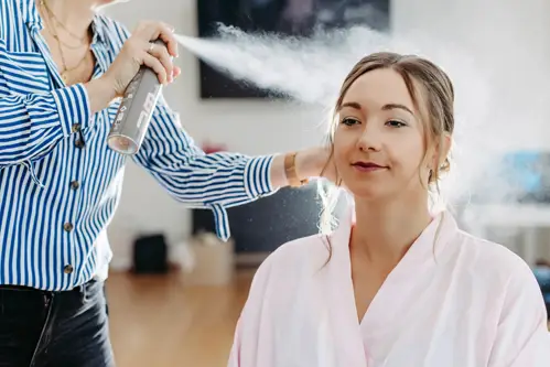 Eine Frau im rosa Morgenmantel lässt sich von einer Person in blau-weiß gestreiftem Hemd die Haare stylen und sprüht dabei gekonnt Haarspray auf. In dem hellen Raum, in dem eine entspannte und konzentrierte Atmosphäre herrscht.