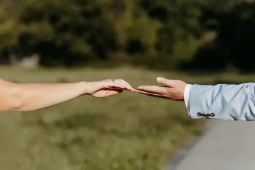 Zwei Hände, die sich einander entgegenstrecken, sich fast berühren, erzählen eine Geschichte der Verbundenheit. Eine Hand ist sanft ausgestreckt, die andere trägt einen hellen Anzug. Vor dem Hintergrund des verschwommenen Grüns könnte dies ein zeitloser Moment sein, eingefangen von einem Hochzeitsfotografen in Marcolinis Welt.