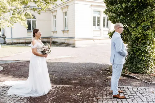 Eine Braut im weißen Kleid mit einem Blumenstrauß in der Hand nähert sich ihrem Bräutigam im hellgrauen Anzug. Sie stehen draußen auf einem Kopfsteinpflasterweg in der Nähe eines prächtigen Gebäudes, umgeben von üppigem Grün. Ein Hochzeitsfotograf in Marcolinis Welt hat das Bild wunderschön festgehalten.