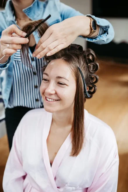 Eine lächelnde Frau in einem rosa Morgenmantel lässt sich die Haare frisieren, möglicherweise für ein Shooting mit einem Hochzeitsfotografen in Marcolinis Welt. Ein Stylist teilt ihr Haar mit Lockenwicklern und hält einen Kamm in der Hand. Im Hintergrund ist eine Inneneinrichtung mit Holzboden zu sehen.