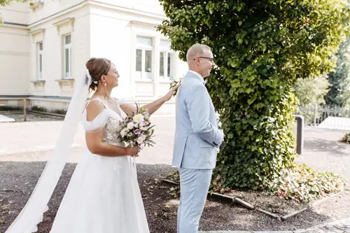 In der bezaubernden Kulisse von Marcolinis Welt berührt eine Braut im weißen Kleid zart die Schulter ihres Bräutigams, der einen hellblauen Anzug trägt. Vor der Kulisse eines üppigen Baumes und eines eleganten Gebäudes wird dieser Moment von einem Hochzeitsfotografen wundervoll festgehalten.