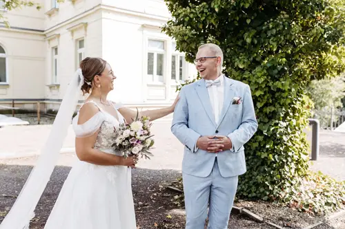 Eine Braut im weißen Kleid hält einen Blumenstrauß in der Hand und lächelt ihrem Bräutigam im hellblauen Anzug unter einem prächtigen Baum entgegen. Ein Hochzeitsfotograf in Marcolinis Welt hält sie fest und teilt einen emotionalen und freudigen Moment vor der eleganten Kulisse des hellen Gebäudes.