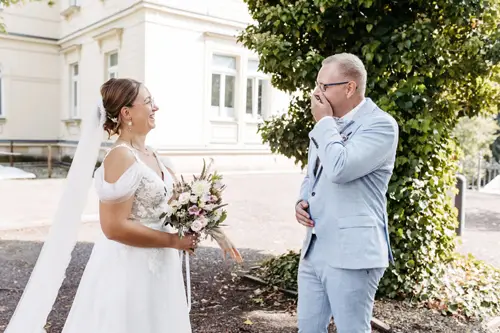 Eine Braut im weißen Kleid hält einen Blumenstrauß in der Hand und lächelt ihrem Bräutigam im hellblauen Anzug unter einem prächtigen Baum entgegen. Ein Hochzeitsfotograf in Marcolinis Welt hält sie fest und teilt einen emotionalen und freudigen Moment vor der eleganten Kulisse des hellen Gebäudes.