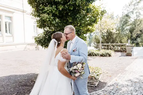 Braut und Bräutigam küssen sich zart, eingefangen von einem Hochzeitsfotografen in Marcolinis Welt. Die Braut, strahlend in einem schulterfreien Kleid und Schleier, hält ihren Brautstrauß, während der Bräutigam sie in einem hellblauen Anzug begleitet. Sie stehen auf einem Kopfsteinpflasterweg, umgeben von üppigem Grün.