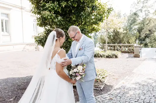 Braut und Bräutigam lächeln sich im Freien freudig an, eingefangen von einem Hochzeitsfotografen in Marcolinis Welt. Die Braut, strahlend in einem schulterfreien Kleid und Schleier, hält ihren Brautstrauß, während der Bräutigam sie in einem hellblauen Anzug begleitet. Sie stehen auf einem Kopfsteinpflasterweg, umgeben von üppigem Grün.