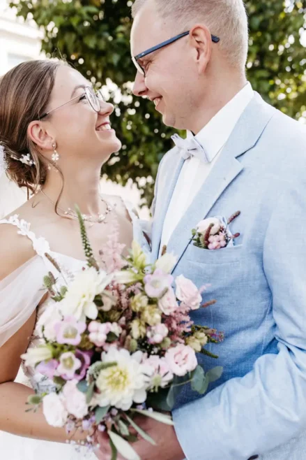 Ein talentierter Hochzeitsfotograf in Marcolinis Welt hält ein Paar fest, das sich an seinem Hochzeitstag liebevoll umarmt. Die Braut in einem floralen Spitzenkleid hält einen Strauß rosa und weißer Blumen in der Hand, während sie sich unter dem sonnendurchfluteten Baum herzlich anlächeln. Der Bräutigam trägt einen hellblauen Anzug.