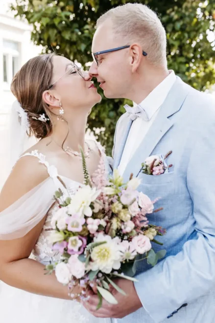 Ein talentierter Hochzeitsfotograf in Marcolinis Welt hält ein Paar fest, das sich an seinem Hochzeitstag liebevoll umarmt. Die Braut in einem floralen Spitzenkleid hält einen Strauß rosa und weißer Blumen in der Hand, während sie sich unter dem sonnendurchfluteten Baum herzlich anlächeln. Der Bräutigam trägt einen hellblauen Anzug.