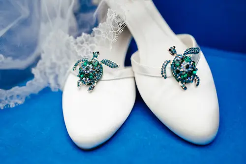 Weiße Brautschuhe, deren dekorative Schildkrötenbroschen mit blauen und grünen Edelsteinen das Licht einfangen, ruhen elegant auf einer leuchtend blauen Oberfläche. Zarter Spitzenstoff blitzt im Hintergrund hervor – eine perfekte Szene für jeden Hochzeitsfotografen in Marcolinis Welt.
