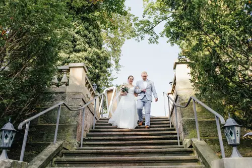 Braut und Bräutigam steigen eine Steintreppe in Marcolinis Welt hinab, umgeben von üppigem Grün. Die Braut im weißen Kleid mit dem Brautstrauß in der Hand und der Bräutigam im grauen Anzug sind perfekte Motive für jeden Hochzeitsfotografen, während Laternen ihren Weg unter sonnendurchfluteten Bäumen sanft erhellen.