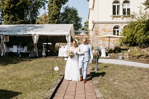 Braut und Bräutigam gehen lächelnd Arm in Arm einen Gartenweg entlang. Die Braut trägt ein weißes Kleid mit Blumenstrauß, der Bräutigam einen hellblauen Anzug. Weiße Stühle und Laternen schmücken die Rasenfläche, dahinter steht ein Zelt – wunderschön eingefangen von Melanie Kunert Fotografie, Hochzeitsfotografin Dresden.