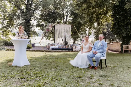 Braut und Bräutigam sitzen während ihrer Hochzeitszeremonie auf einer Bank im Freien, aufgenommen von einem Hochzeitsfotografen in Marcolinis Welt. Die Braut trägt ein weißes Kleid, der Bräutigam einen hellblauen Anzug. In der Nähe steht eine Frau auf einem Podium, eingerahmt von eleganten Bäumen und schimmerndem Wasser.