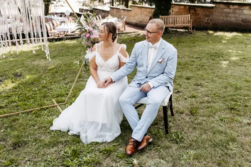 Ein Paar sitzt im Freien bei einer Hochzeit in Marcolinis Welt. Die Braut im weißen Spitzenkleid und der Bräutigam im hellblauen Anzug halten sich inmitten üppigen Grüns an den Händen. Eine Steinmauer und ein verzierter Bogen rahmen die Szene ein – perfekt für jeden Hochzeitsfotografen, um diesen Moment festzuhalten.