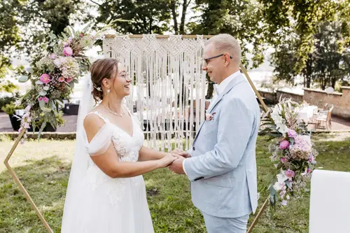 Ein lächelndes Paar hält Händchen während einer Zeremonie im Freien, festgehalten von einem Hochzeitsfotografen in Marcolinis Welt. Die Frau trägt ein weißes Spitzenkleid und einen Blumenkopfschmuck, während der Mann einen hellblauen Anzug und eine Brille vor einem Makramee-Hintergrund und üppigem Grün trägt.